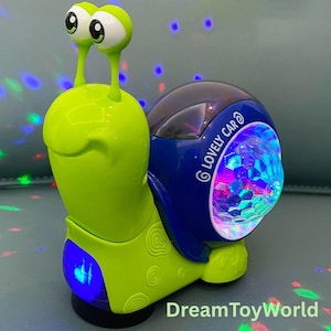 Puede incluir: Un juguete en forma de caracol verde y azul con ojos grandes y una concha que se ilumina. La concha tiene la inscripción "LOVELY CAR". El juguete está diseñado para proyectar luces de colores.