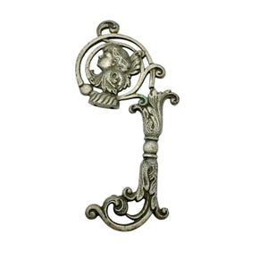 Pode incluir: Peça decorativa em forma de chave de metal prateado ornamentada. O design apresenta um perfil de rosto de querubim dentro de uma moldura circular, com intrincados detalhes florais e de volutas. A chave tem uma estética vintage e antiga.