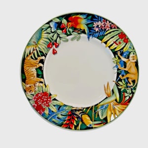 Peut inclure: Une assiette en céramique blanche avec une bordure sur le thème de la jungle. La bordure présente un tigre, un singe, un perroquet et diverses fleurs et feuillages tropicaux verts, bleus, rouges et jaunes. L'assiette a un bord vert.