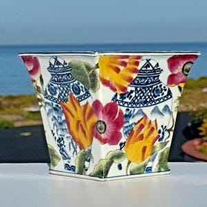 Könnte beinhalten: Ein quadratischer, weißer Keramik-Blumentopf mit einem lebendigen Blumendesign. Der Pflanztopf zeigt rote, gelbe und rosa Blumen sowie blau-weiß gemusterte Vasen. Der Hintergrund zeigt einen Blick auf das Meer.
