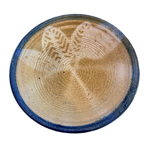 May include: A round ceramic plate featuring a speckled tan and beige center with concentric circular grooves. The plate has a distinct blue glazed rim and a faint, light-colored stamped pattern of two symmetrical leaf-like shapes near the top.