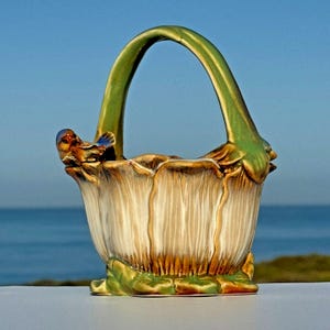 May include: A ceramic basket with a green handle and accents. The basket is shaped like a flower with cream and brown petals. A small bird figurine is perched on the rim. The background is a blurred view of the ocean.