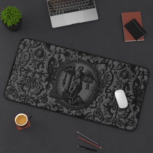 May include: A dark grey rectangular mouse mat with an ornate baroque pattern. The centre features a circular emblem with a classical figure. A white computer mouse sits on the mat, next to a laptop, notebook, and pencils.