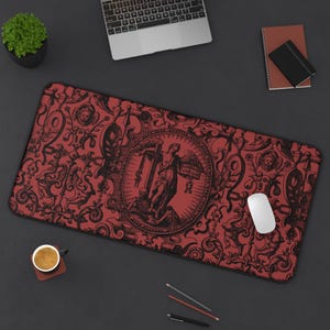 May include: A red and black rectangular desk mat with an ornate, vintage design. The mat features a central circular illustration with a figure and text, surrounded by scrolling patterns and decorative elements. A white mouse rests on the mat.