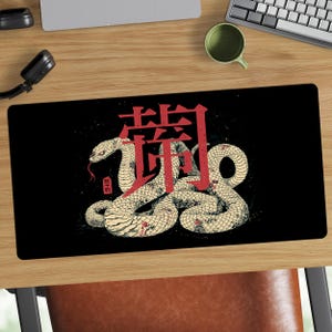 Può includere: Tappetino per mouse rettangolare nero con un'illustrazione dettagliata di un serpente color crema con accenti rossi e un carattere giapponese rosso. Il serpente è arrotolato e lo sfondo è nero.