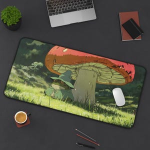 May include: A rectangular mouse pad featuring a whimsical illustration of a frog wearing a hat and cloak, sheltering under a large mushroom. The scene is set in a grassy field with a forest background. The pad is black with a white mouse and a cup of coffee.