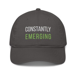 May include: A dark gray baseball cap with the words "CONSTANTLY EMERGING" embroidered in white and green thread. The cap has a curved brim and a structured fit, suitable for casual wear.