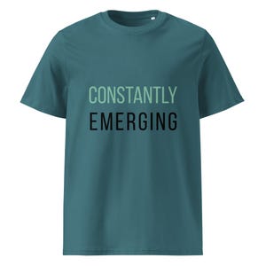 May include: Teal-colored t-shirt with the words "CONSTANTLY EMERGING" printed on the front. The top text is in a light green color, and the bottom text is in black. The shirt has short sleeves and a crew neck.