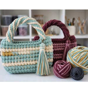 Pode incluir: Duas bolsas de crochê feitas à mão em verde-azulado e bordô, com borlas a condizer. A bolsa verde-azulado tem um padrão listrado creme e amarelo e uma alça trançada. Também são visíveis novelos de lã em cores coordenadas, sugerindo um projeto artesanal.