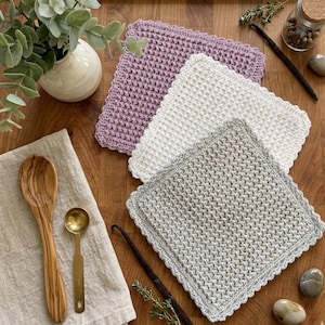 May include: A collection of four square, textured washcloths in shades of lavender, white, and gray, arranged on a wooden surface. A wooden spoon, measuring spoon, and vanilla beans are also present. The washcloths have a scalloped edge.