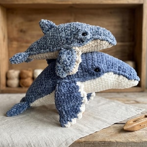 May include: Two plush whales, one atop the other, in shades of blue and white. The whales are handmade, with a textured, soft appearance. The larger whale is a darker blue, while the smaller whale is a lighter blue. The underside of both whales is white.