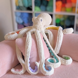 Puede incluir: Un pulpo blanco de peluche hecho a mano con puntas de tentáculos de colores del arcoíris descansa sobre un cojín de pana rosa. El pulpo tiene un aspecto texturizado y tejido. El fondo presenta una estantería llena de hilo colorido.