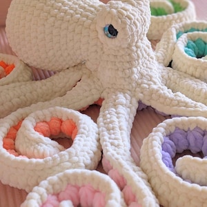 Könnte beinhalten: Ein handgefertigtes Kuscheltier in Form eines Oktopus aus cremefarbenem Garn. Der Oktopus hat blaue Augen und Tentakel mit farbigen Akzenten in Rosa, Orange, Grün und Lila. Das Spielzeug ist weich und kuschelig.