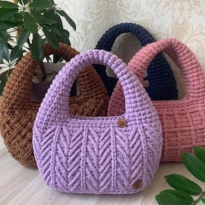 May include: Four handmade crochet handbags in shades of brown, lavender, navy, and pink. The bags have rounded shapes with thick handles. The lavender bag features a chevron pattern, while the others have textured designs. Each bag has a small, round, brown tag.
