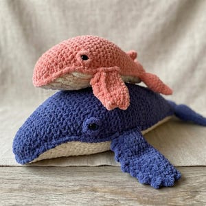 May include: Two crocheted whale plushies. The larger whale is a deep blue, and the smaller whale is a soft pink. Both have black eyes and are made of a textured yarn. The whales are resting on a neutral-coloured surface.