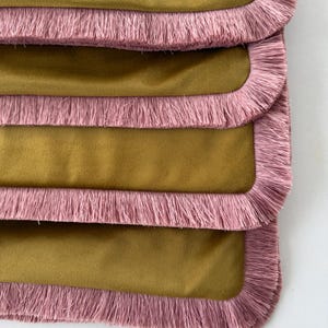 May include: A stack of rectangular fabric pieces in a muted olive green colour. Each piece features a soft, velvet-like texture and is finished with a thick, light pink fringe trim that runs along all four rounded edges of the fabric.