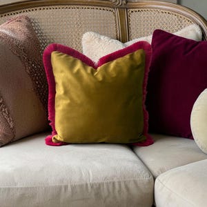 Könnte beinhalten: Eine Sammlung dekorativer Kissen in verschiedenen Farben und Texturen auf einem cremefarbenen Sofa. Die Kissen umfassen ein senfgelbes Samtkissen mit fuchsiafarbener Fransen, ein bordeauxfarbenes Samtkissen und ein strukturiertes rosa Kissen.