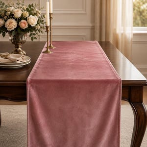 Könnte beinhalten: Ein rechteckiger Tischläufer aus altrosa Samtstoff mit einer glatten, weichen Textur und gesäumten Kanten. Der Läufer liegt flach in der Mitte eines dunklen Holztisches und zeichnet sich durch eine gleichmäßige Farbe und matte Oberfläche aus.