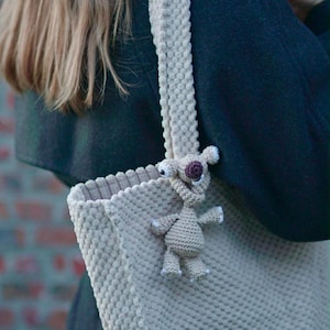 Pode incluir: Uma sacola tote bege de crochê com uma alça combinando. Uma pequena criatura de crochê com uma flor roxa na cabeça está presa à bolsa. A bolsa tem um padrão texturizado em relevo.