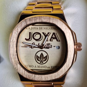 May include: Rose gold wristwatch with a rectangular face. The watch face features the word "JOYA" and other text. The watch is presented in a yellow box with a white pillow.