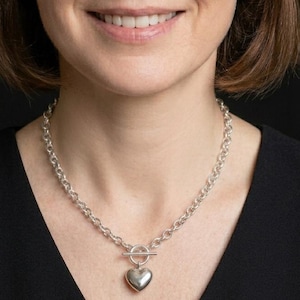 Può includere: Una collana a catena color argento con chiusura a T sul davanti. Un ciondolo a forma di cuore, liscio e lucido, pende dal centro della barra di chiusura, creando un classico design a maglie con una finitura metallica.