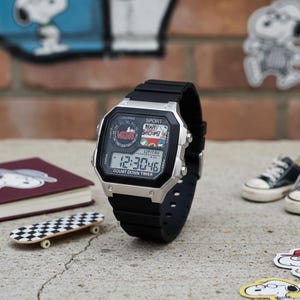 Könnte beinhalten: Schwarze und silberne Digitaluhr mit schwarzem Armband. Das Zifferblatt zeigt die Uhrzeit, Snoopy-Grafiken und den Text "Vans" und "Sport". Die Uhr liegt auf einer Betonoberfläche.