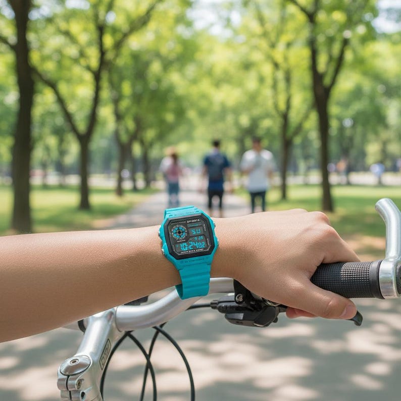 May include: A turquoise digital watch with a black face is worn on a wrist, with the watch displaying the time. The watch is on a person's wrist, holding a bicycle handlebar. The background shows a park with people walking.
