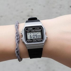 以下が含まれることがあります： 黒いバンドが付いたシルバーのCasio F-91Wデジタル時計は、6:09:00の時刻を表示しています。時計は、灰色の編み込みブレスレットの隣の腕にあります。時計には、Casioのロゴが付いた長方形の文字盤があります。