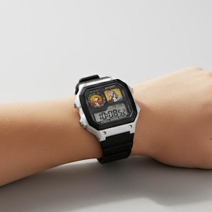 May include: A black and white digital watch with a black band is worn on a wrist. The watch face displays the time and features cartoon character graphics. The watch reads "SPORT" at the top.