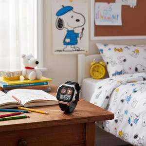 May include: A black and silver digital watch with a Snoopy graphic on the face. The watch is displayed on a wooden table next to an open book and colored pencils. A Snoopy drawing hangs on the wall in the background.