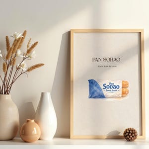 Puede incluir: Una impresi&oacute;n enmarcada con el texto "PAN SOBAO" y una ilustraci&oacute;n de pan dulce. La impresi&oacute;n est&aacute; en un marco de madera clara y se muestra sobre una superficie blanca. Hay jarrones decorativos y flores a la izquierda.