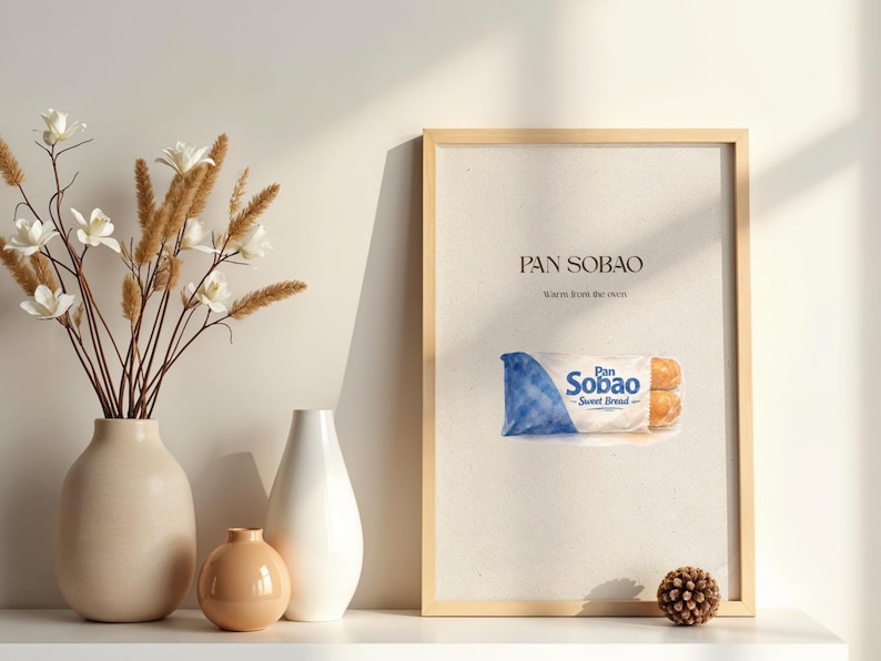 Puede incluir: Una impresi&oacute;n enmarcada con el texto "PAN SOBAO" y una ilustraci&oacute;n de pan dulce. La impresi&oacute;n est&aacute; en un marco de madera clara y se muestra sobre una superficie blanca. Hay jarrones decorativos y flores a la izquierda.
