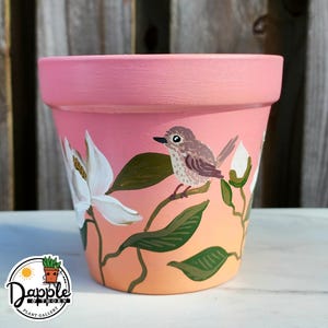 May include: A hand-painted terracotta flower pot with a pink rim and a gradient peach and pink body. The pot features a white magnolia flower and a small bird perched on a branch. The pot has the logo of Dapple & Thorn Plant Gallery.