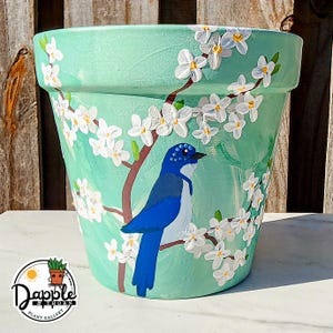 Puede incluir: Maceta de cerámica turquesa pintada a mano con un pájaro azul posado en una rama floreciente. Flores blancas con centros amarillos y ramas marrones adornan la maceta. La maceta tiene un logotipo redondo que dice "Dapple & Thorn Plant Gallery".