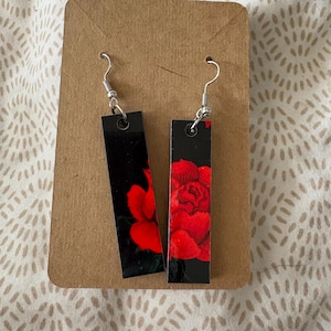 May include: A pair of rectangular earrings with a black background and a red rose design. The earrings are displayed on a brown card with silver-colored ear wires.