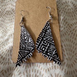 Puede incluir: Un par de pendientes estampados en blanco y negro con un diseño geométrico. Los pendientes tienen forma triangular y cuelgan de ganchos plateados. Los pendientes se exhiben en una tarjeta marrón.