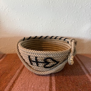 May include: A round bowl constructed from coiled tan rope, featuring a black stitched heart and arrow design on the exterior. The upper rim is accented with black stitching, and a decorative knotted rope detail is attached to the side.