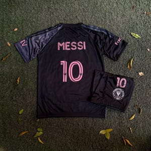 May include: A black soccer jersey and matching shorts set featuring the name MESSI and the number 10 in pink lettering. The jersey has short sleeves with dark stripes, and the shorts display the number 10 and the Club Internacional de Futbol Miami logo.