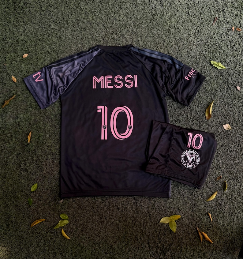 May include: A black soccer jersey and matching shorts set featuring the name MESSI and the number 10 in pink lettering. The jersey has short sleeves with dark stripes, and the shorts display the number 10 and the Club Internacional de Futbol Miami logo.