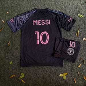 May include: A black soccer jersey and matching shorts set featuring the name MESSI and the number 10 in pink lettering. The jersey has dark grey sleeves with the text tv and Frac on the shoulders. The shorts display the number 10 and a circular logo.