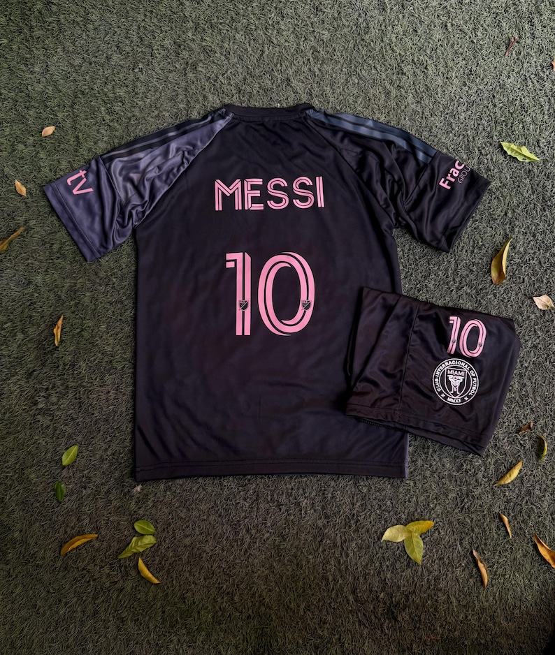 May include: A black soccer jersey and matching shorts set featuring the name MESSI and the number 10 in pink lettering. The jersey has dark grey sleeves with the text tv and Frac on the shoulders. The shorts display the number 10 and a circular logo.