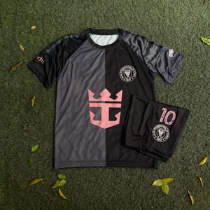 May include: A short-sleeved athletic shirt and matching shorts set in a split black and dark gray color scheme. The shirt features a large pink crown logo on the front, a circular Miami soccer club crest, and a small logo on the right sleeve.