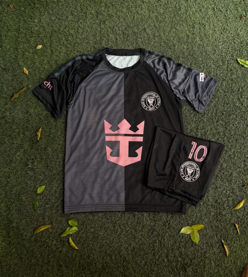May include: A short-sleeved athletic shirt and matching shorts set in a split black and dark gray color scheme. The shirt features a large pink crown logo on the front, a circular Miami soccer club crest, and a small logo on the right sleeve.