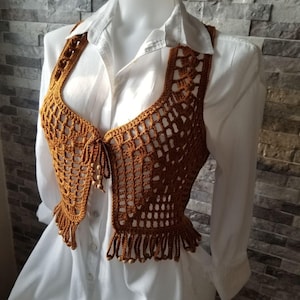 Peut inclure: Un gilet en crochet marron avec une bordure à franges et un motif ajouré décoratif. Le gilet est présenté sur une chemise blanche boutonnée sur un mannequin. Le gilet a une encolure dégagée et est orné de petites perles en bois.