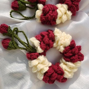 Peut inclure: Deux chouchous faits à la main au crochet. Chaque chouchou est un anneau de mailles au crochet crème et bordeaux alternées. Des tiges vertes avec des bourgeons au crochet bordeaux sont attachées à chaque chouchou. Les chouchous sont sur une surface en satin blanc.