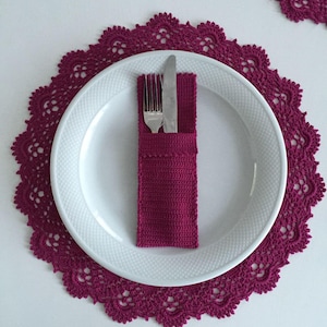 May include: A white plate with a fork and knife in a magenta crochet holder, set on a matching magenta crochet placemat. The placemat has a scalloped edge. The image is taken from above.