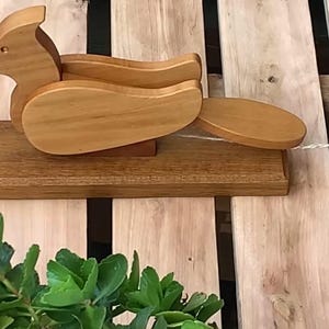 May include: A wooden pull-along duck toy with a light brown finish. The duck has a long neck and a rounded body, attached to a rectangular base. A string extends from the duck for pulling. The toy is on a wooden surface.