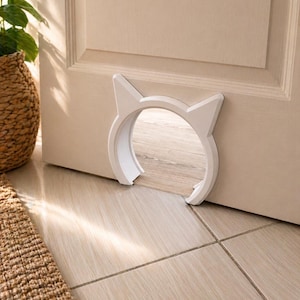 Peut inclure: Une chatière blanche en forme de chat installée dans une porte beige. La porte est découpée en forme circulaire avec deux oreilles pointues en haut. La porte est installée au-dessus d'un carrelage clair.