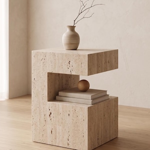May include: A beige stone side table with a unique, open design. A small, beige vase with a twig sits on top. Below, a stack of books and a wooden sphere are displayed in the open space. The table is set on a wooden floor.