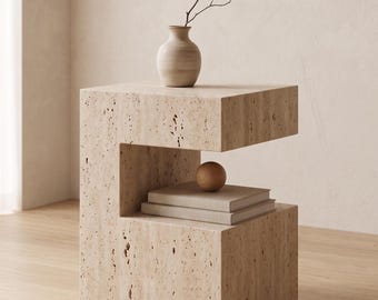 May include: A beige stone side table with a unique, open design. A small, beige vase with a twig sits on top. Below, a stack of books and a wooden sphere are displayed in the open space. The table is set on a wooden floor.